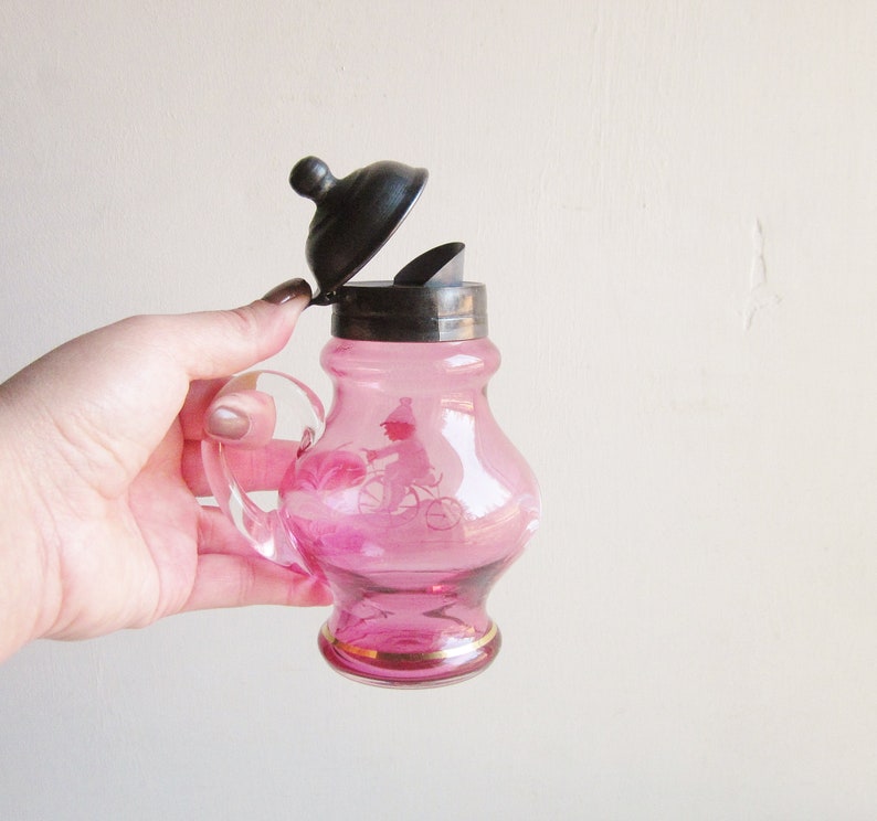 May include: A pink glass creamer with a silver lid and a bicycle design. The creamer has a gold rim.