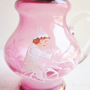 May include: A pink glass pitcher with a clear handle. The pitcher has a hand-painted design of a person riding a bicycle.