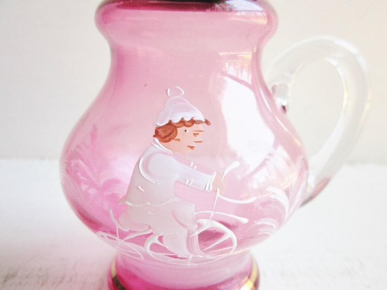 May include: A pink glass pitcher with a clear handle. The pitcher has a hand-painted design of a person riding a bicycle.