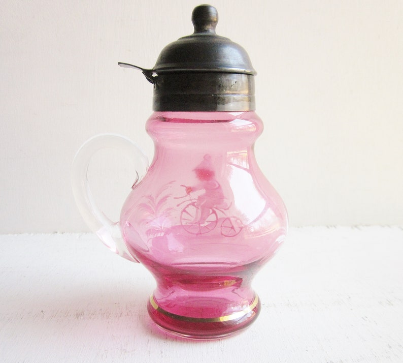 May include: A pink glass creamer with a silver lid and a handle. The creamer has a gold trim around the bottom and a design of a person riding a bicycle.