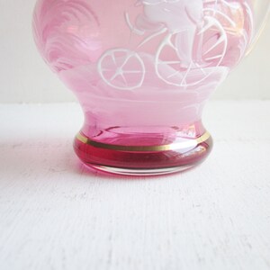 May include: A pink glass pitcher with a white etched design of a person riding a bicycle. The pitcher has a gold rim around the base.