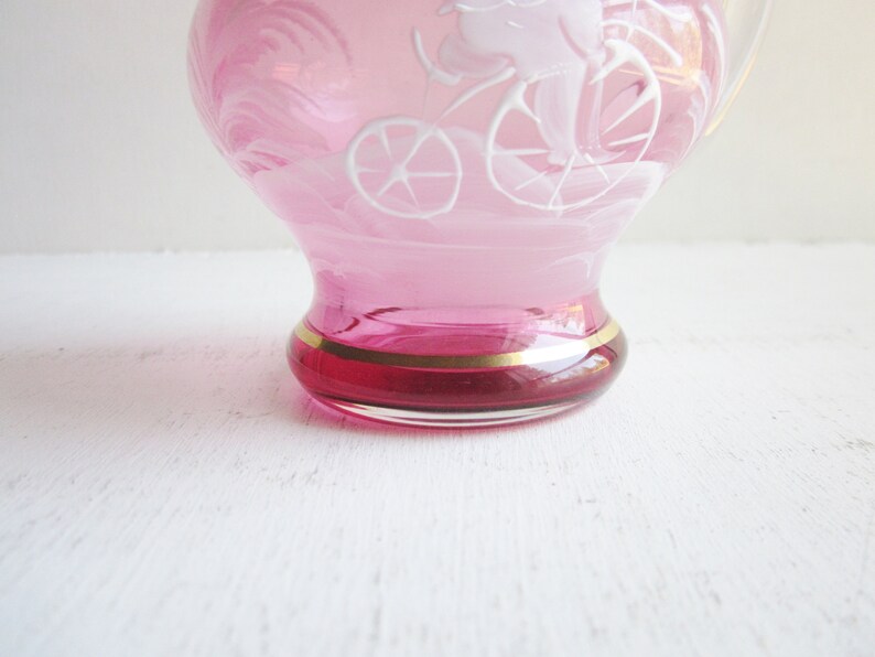 May include: A pink glass pitcher with a white etched design of a person riding a bicycle. The pitcher has a gold rim around the base.