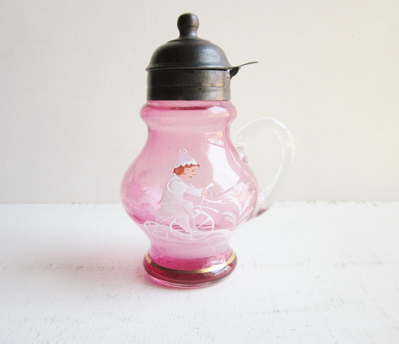 May include: A pink glass creamer with a silver lid and a painted design of a person riding a bicycle. The creamer has a gold trim around the base.
