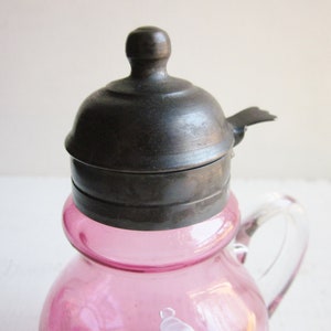 May include: A pink glass creamer with a silver lid and a clear glass handle. The lid has a small spout on the side.