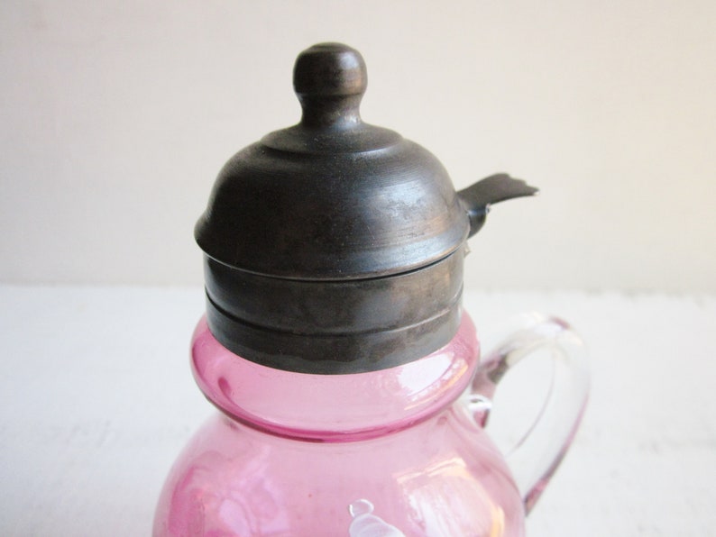 May include: A pink glass creamer with a silver lid and a clear glass handle. The lid has a small spout on the side.