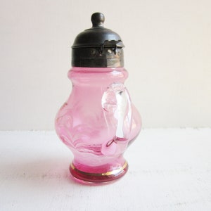 May include: A pink glass pitcher with a silver lid and a clear glass handle. The pitcher has a gold rim and a swirl design.