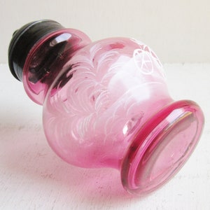 May include: A pink glass bottle with a black lid and white etched designs. The bottle has a round body and a wide mouth.