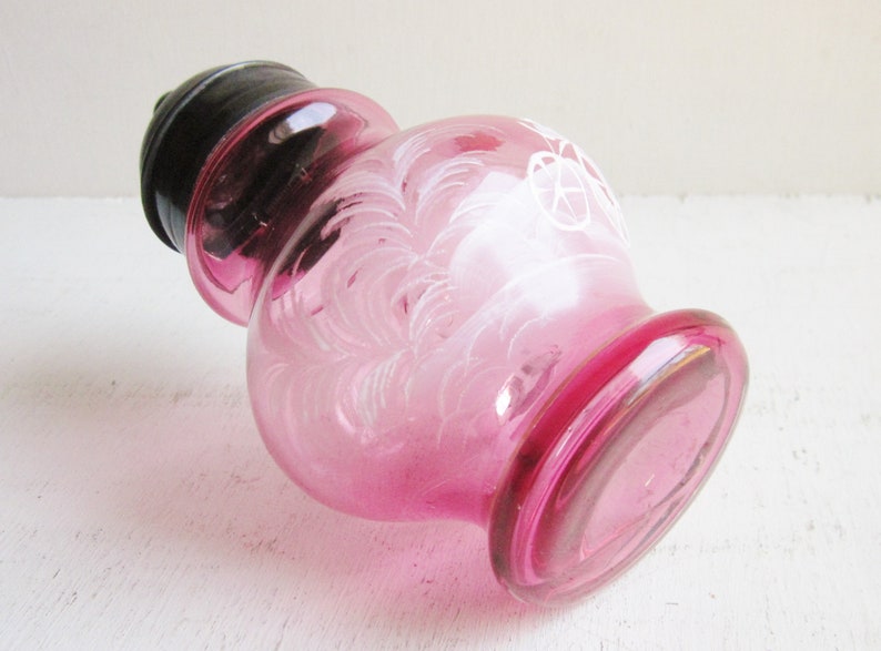 May include: A pink glass bottle with a black lid and white etched designs. The bottle has a round body and a wide mouth.