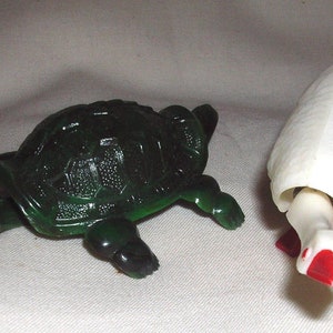 Könnte beinhalten: Zwei Plastik-Tierfiguren: eine dunkelgrüne Schildkröte und eine weiße Gans. Die Schildkröte hat einen detaillierten Panzer und sichtbare Beine. Die Gans ist nach unten gebeugt mit roten Füßen und Schnabel.