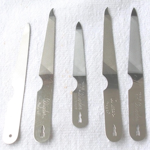 May include: A collection of nail files in various sizes. The files are made of metal and have a textured surface for filing nails. Some files have the brand name "Wiader" engraved on them. The files are arranged on a white surface.