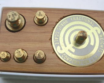 Puede incluir: Un juego de pesas fraccionarias Ohaus vintage. El juego incluye pesas de latón de varios tamaños, un dial circular y una base de madera. Las pesas están dispuestas en una bandeja de plástico blanco. El dial es dorado con el texto "Ohaus Fractional Weights Corporation U.S.A."