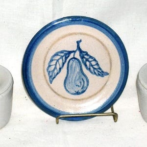 May include: A ceramic set featuring two lidded mugs and a small plate. The plate and mug lids have a dark blue accent. The plate has a blue pear design. The set is white with blue accents.