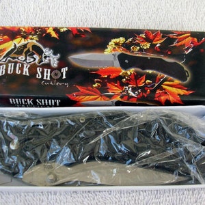 Könnte beinhalten: Ein schwarzes Klappmesser mit einer silbernen Klinge in einer weißen Schachtel mit einem roten, orangefarbenen und gelben Herbstlaubdesign. Die Schachtel trägt den Text "Buck Shot Cutlery" und "Buck Shot".