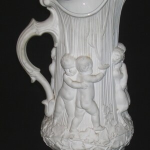 May include: White ceramic pitcher with a detailed relief design. The pitcher features a handle and a spout. The body is adorned with cherubic figures and vertical textured lines, with a base of leaves.