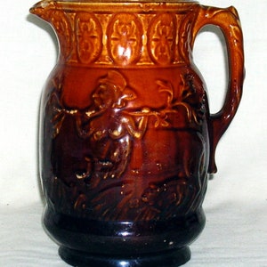 Pode incluir: Um jarro de cerâmica marrom com uma alça e um desenho em relevo de uma mulher segurando um galho. O jarro tem um esmalte marrom escuro e uma superfície texturizada.