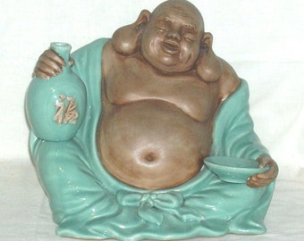 Può includere: Una statuetta in ceramica di un Buddha sorridente con la pancia rotonda che tiene una bottiglia verde e una piccola ciotola. Il Buddha indossa una tunica turchese e ha grandi orecchie carnose.