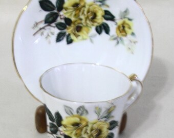 May include: A white teacup and saucer set with a gold rim and a floral design featuring yellow roses and green leaves.