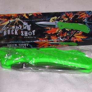 Könnte beinhalten: Ein grünes Taschenmesser mit schwarzem Griff und silberner Klinge. Das Messer ist in einer Plastiktüte verpackt und hat eine schwarze Schachtel mit der Aufschrift "Buck Shot Cutlery".