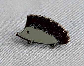 Puede incluir: Un pin de esmalte de erizo de color plateado. El pin presenta un cuerpo gris claro, una espalda espinosa marrón oscuro y un pequeño ojo negro. El erizo tiene cuatro patas pequeñas y es un accesorio lindo.