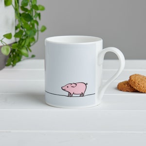 May include: A white ceramic mug with a light blue background and a pink pig illustration. The pig is walking to the right and has a curled tail.