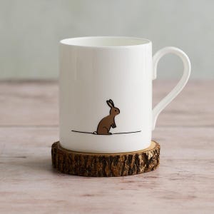 May include: White ceramic mug with a brown rabbit illustration. The mug sits on a wooden coaster with a natural bark edge. The handle is a curved, loop shape.