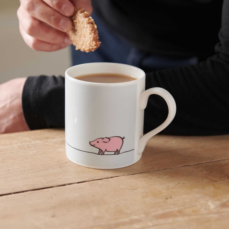 May include: A white ceramic mug with a pink pig illustration and a black outline. The mug is filled with a brown liquid and has a black handle.