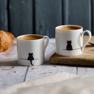 Puede incluir: Dos tazas de cerámica blancas llenas de café. Cada taza presenta una silueta de gato negro sentado en una línea. Las tazas están sobre una superficie de madera, con un cruasán visible en el fondo. Diseño sencillo y moderno.