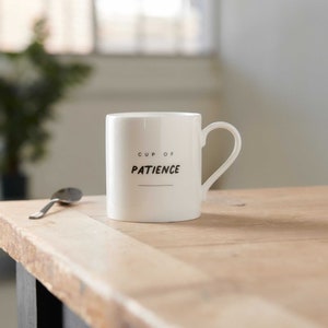 May include: White ceramic mug with a black inscription that reads "CUP OF PATIENCE". The mug is sitting on a wooden table.
