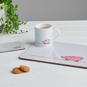 May include: A white ceramic mug with a pink pig illustration and a gray coaster with a pink pig illustration. The mug and coaster are on a white wooden table with two cookies.