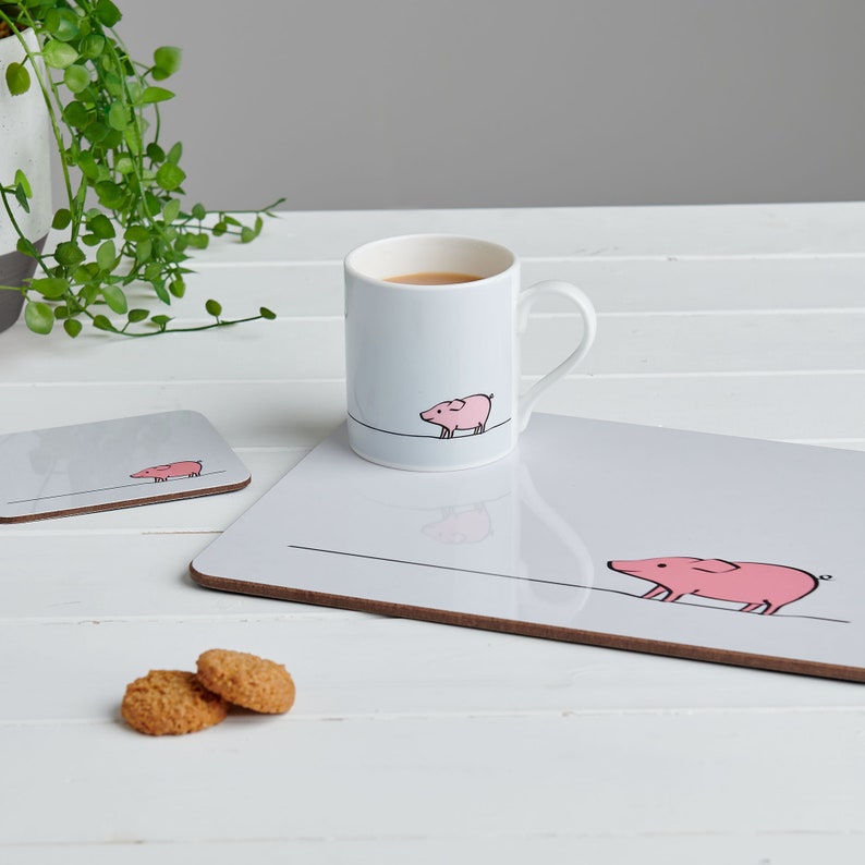 May include: A white ceramic mug with a pink pig illustration and a gray coaster with a pink pig illustration. The mug and coaster are on a white wooden table with two cookies.