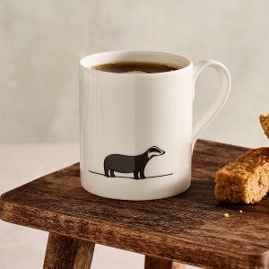 May include: A white ceramic mug filled with a dark liquid, featuring a black and gray badger illustration. The mug sits on a wooden surface, with a handle on the right side. A piece of baked good is on the right.