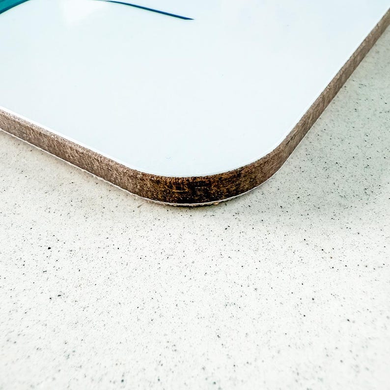 May include: A white, rectangular coaster with a brown, wood-like edge. The coaster has a glossy finish and a subtle design in the upper left corner. The coaster is set against a speckled, light-colored surface.