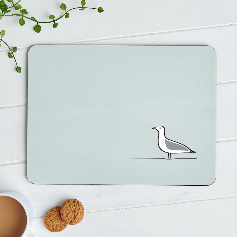 May include: A light blue rectangular mousepad with a minimalist illustration of a seagull. The seagull is depicted in black and white with a yellow beak, standing on a thin black line. The mousepad is on a white wooden surface.