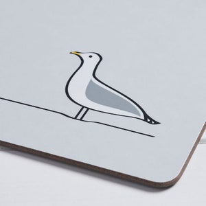 May include: A rectangular coaster featuring a stylized illustration of a seagull perched on a line. The seagull is white and gray with black outlines and a yellow beak. The coaster has a light gray background and a brown edge.