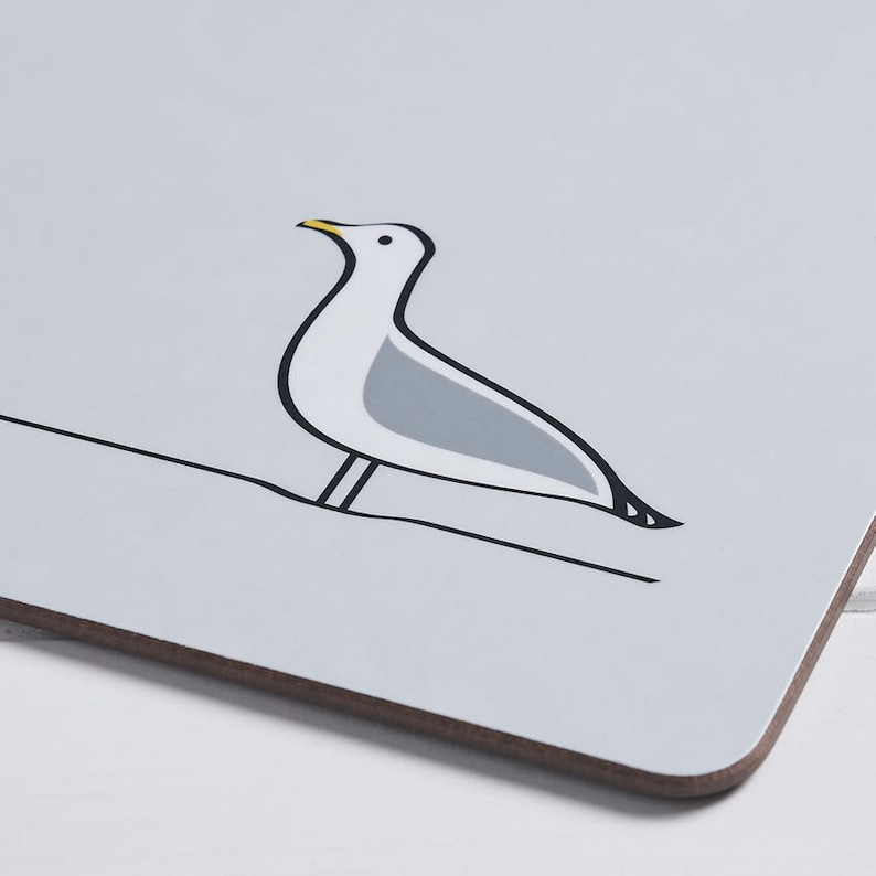 May include: A rectangular coaster featuring a stylized illustration of a seagull perched on a line. The seagull is white and gray with black outlines and a yellow beak. The coaster has a light gray background and a brown edge.