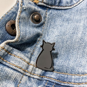 Könnte beinhalten: Eine silberne Metallstifte mit der Silhouette einer schwarzen Katze auf einem blauen Jeansstoff.