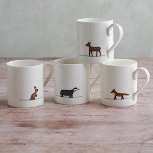 May include: Four white ceramic mugs with curved handles. Each mug features a different animal illustration: a brown deer, a brown rabbit, a black badger, and a red fox. The animals are depicted in a minimalist style.