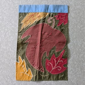 May include: A rectangular garden flag featuring an olive green background with a light blue top border. The design includes appliqued fabric leaves in shades of mustard yellow, deep red, and maroon, accented with decorative white stitching.