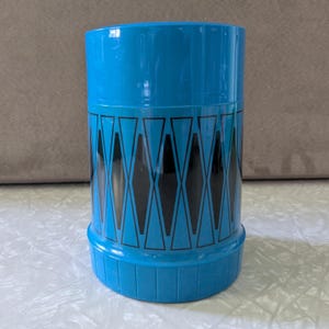 May include: A cylindrical bright blue plastic container featuring a repeating black geometric diamond pattern around the center. The base of the container has a series of vertical ridges, and the top section is a smooth, solid blue finish.