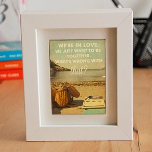 May include: A white picture frame with a vintage-style print inside. The print features a beach scene with a basket, sandals, and a record player. The text on the print reads "We're in love. We just want to be together. What's wrong with that?"