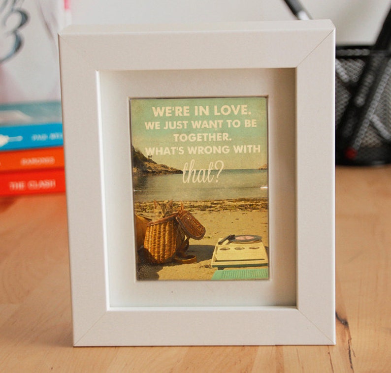 May include: A white picture frame with a vintage-style print inside. The print features a beach scene with a basket, sandals, and a record player. The text on the print reads "We're in love. We just want to be together. What's wrong with that?"