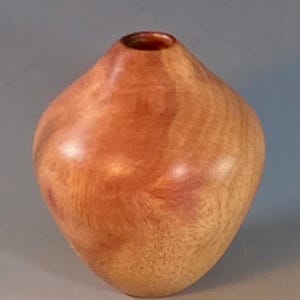 May include: A handcrafted wooden vase with a rounded, diamond-like shape. The vase is made of light brown wood with reddish-brown accents and a smooth, polished finish. The opening is small and circular.