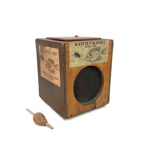 May include: A vintage wooden Katch-N-Karry Roach Trap with a mesh front and a small wooden stopper. The trap has two labels with illustrations of a fish and a bug. The text "Katch-N-Karry Roach Trap" is printed on the labels.