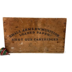 May include: A vintage wooden crate with the text "SMALL ARMS AMMUNITION LOADED PAPER SHOT GUN CARTRIDGES" printed in black. A colorful spinning top with red, green, and yellow stripes sits in front of the crate. The crate is made of aged wood.