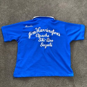 May include: A blue short-sleeved polo shirt with a white collar and white lettering on the back that reads "Jim Harrington's Apache Ski-Doo Suzuki".