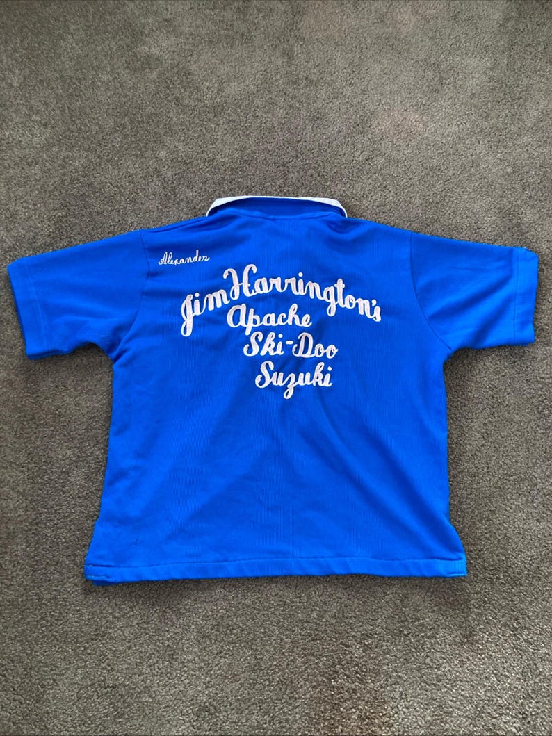 May include: A blue short-sleeved polo shirt with a white collar and white lettering on the back that reads "Jim Harrington's Apache Ski-Doo Suzuki".