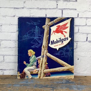 May include: Vintage Mobilgas sign featuring a red Pegasus logo. The sign depicts a woman and a man sitting on a wooden fence, with a blue background showing an airplane and industrial structures. The sign reads "Mobilgas" and "SOCONY-VACUUM".