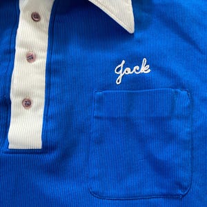 May include: A blue collared shirt with a white collar and a white stripe down the front. The shirt has a pocket on the left side with the word "Jack" embroidered in white.