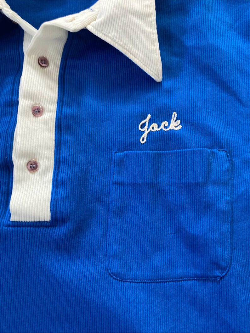May include: A blue collared shirt with a white collar and a white stripe down the front. The shirt has a pocket on the left side with the word "Jack" embroidered in white.