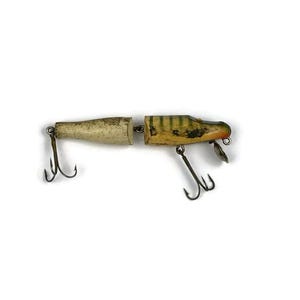 May include: Vintage two-piece fishing lure. The lure is primarily cream-colored with green and yellow stripes and black accents. It features two treble hooks and a small metal fin. The lure is designed to attract fish.